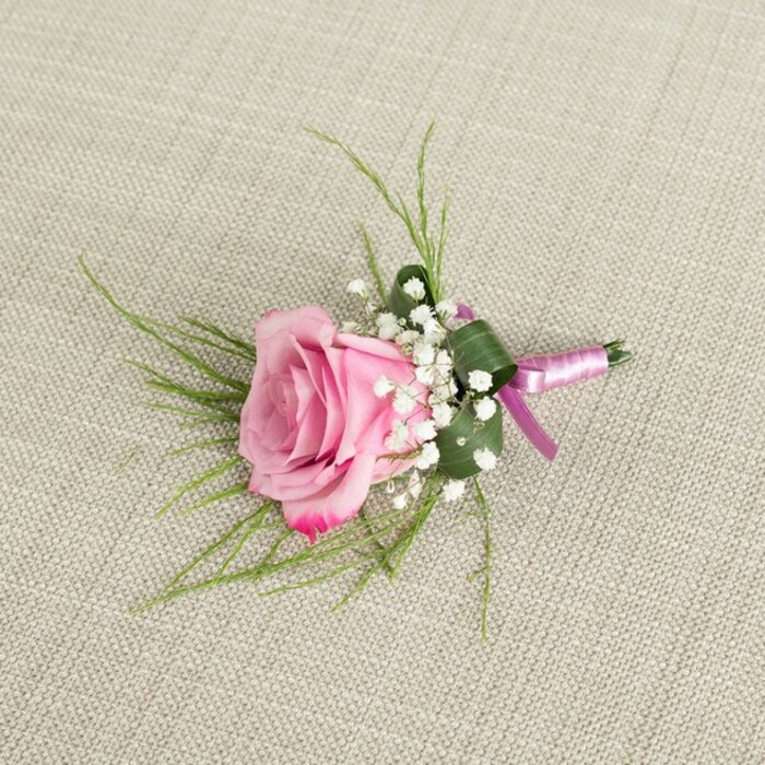 An exquisitely detailed pink rose boutonniere is photographed resting on a pale beige, linen-like fabric, the textured background echoing the understated elegance of a Clerkenwell wedding venue or chic town hall ceremony. The image is captured from an overhead perspective, allowing a full appreciation of the rose's form: its petals are tightly layered near the center, where the pink is almost pastel and powder-soft, gradually unfurling to reveal deeper, more saturated pink at the tips, with a hint of magenta at the very edges. The rose is surrounded at its base by generous tufts of white baby's breath, each tiny bloom clustered together to form a soft, clouded cushion that contrasts beautifully with the rose's bold structure. Emerging from behind the focal flower, slender, grass-like green foliage radiates gently outward, introducing a light, feathery movement, while a broader, darker green leaf has been carefully folded and wrapped around the stems to create a snug, tailored cradle, giving the piece a polished, couture finish. The stems themselves are meticulously bound in a slim band of satin ribbon, its surface shimmering in tones of light lavender-pink melting into a richer magenta, secured in a compact, tidy knot at the base. Subtle, diffused lighting casts mild shadows that bring out the linen's weave and the three-dimensional quality of each petal and sprig. The overall impression is one of careful craftsmanship and quiet romance, an ideal boutonniere or small corsage for a groom, groomsman, or special guest attending a celebration near Farringdon Station, Spa Fields, or one of Clerkenwell's intimate restaurants, where contemporary city style meets the soft charm of classic flowers.