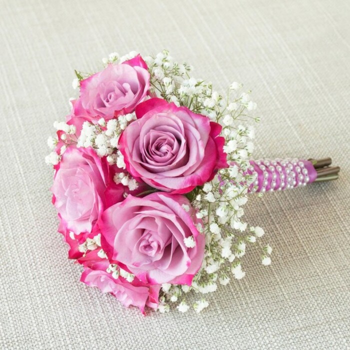 A striking floral creation, this Clerkenwell bouquet draws the eye with its harmonious blend of colour and texture: radiant fuchsia-edged roses cradle softly lavender interiors, the multi-tonal petals reminiscent of sunsets above the city. Five prominent blooms, their forms meticulously arranged, are interlaced with delicate white baby's breath that seems to float above the flowers, giving a gentle, frothy lift to the ensemble. The stems are tightly bound, wrapped with a smooth, rich purple satin ribbon-spiralled with rows of luminous, iridescent pearls that catch the light, creating a sophisticated yet inviting handle. From underneath, hints of fresh green and earth-toned stems ground the bouquet in nature. Bathed in natural light as it rests upon a subtly textured cream linen, this floral piece seems to glow, every detail brought into relief. There is an atmosphere of celebration-a sense of intimacy and delight perfect for marking weddings or special anniversaries in the vibrant Clerkenwell area, whether celebrating at Sadler's Wells or sharing joy in a hidden Rosebery Avenue garden. This bouquet is a testament to the passionate skill and local flair of Clerkenwell's finest florists, inviting new memories with every bloom.