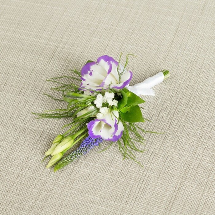 Set against a softly textured light beige fabric, reminiscent of natural linen you might find in a Clerkenwell design studio, this elegant boutonniere has the quiet poise of something handmade with intention. At its centre, two open lisianthus blooms draw the eye: their petals are a clean, luminous white, each delicately crimped at the edges and traced with a band of saturated purple, giving a graceful two-tone effect that feels both classic and contemporary. These focal flowers sit in gentle contrast to the pale background, their rich edges echoing the deep, jewel-like tones often seen in evening weddings around nearby Farringdon and Exmouth Market. Tucked among them are tiny white filler flowers, akin to baby's breath, which add a soft, cloud-like texture and break up the stronger colours with airy lightness. A slim spire of muscari rises beside the lisianthus, its minuscule, bell-shaped blossoms packed closely together in a deep grape-purple that introduces a vertical line and a more textured, almost tactile element. Around the flowers, fine fronds of feathery green foliage, similar to asparagus fern, splay out in light, lacy arcs, offering freshness and a suggestion of movement, as though a breeze had just brushed past. Two small, pale green buds nestle into the arrangement, still closed, hinting at new blooms and adding a gentle note of anticipation. Below, the stems are gathered precisely and bound with a smooth white satin ribbon, firmly wrapped and finished with a modest bow so it can be pinned neatly to a lapel. The overall impression is one of understated sophistication-a carefully curated floral accent ideal for a groom or guest at a city celebration, crafted with the same care you'd expect from an experienced Clerkenwell florist.