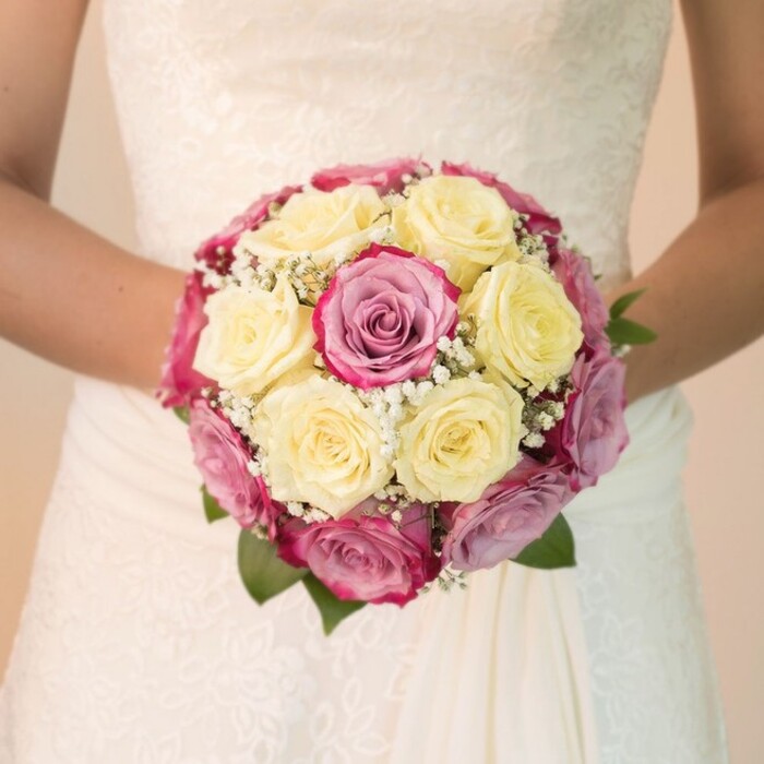 Against the backdrop of a softly lit Clerkenwell wedding venue, a bride stands holding a magnificent, round bouquet close to her waist, her bare arms both protective and proud. Composed mainly of creamy lemon roses and vivid fuchsia pink roses, the bouquet is thoughtfully interspersed with blooms that reveal delicate lavender and dusty mauve as the light shifts across their petals. Tiny white sprays of baby's breath weave through the bouquet, lending an ethereal, cloudlike finish above glossy green leaves that offer a natural frame. The bride's dress, captured from the chest down, features intricate lace that catches the light-its floral motifs dancing along the bodice-before flowing into a skirt of smooth, draping fabric. The lighting is bright yet perfectly balanced, accentuating every texture: the satiny touch of the roses, the gentle frill of baby's breath, and the undulating lacework on the dress. The background is wonderfully understated, softly blurred in gentle tones that place the bouquet-and the care of the local florist-firmly at center stage. This composition radiates anticipation and romance, reflecting both the bespoke craftsmanship found in Clerkenwell and the emotional resonance of a wedding day. It's an ideal image for inspiring couples, celebrating local floral design, or illustrating wedding memories that bloom in the vibrant city heart of Clerkenwell.