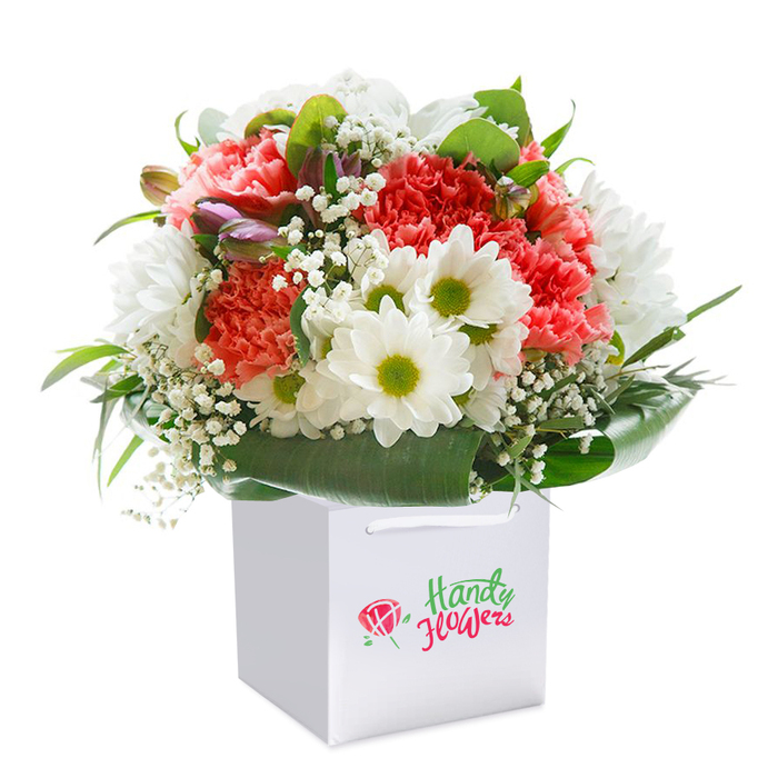 In the heart of Clerkenwell, this vibrant floral bouquet stands out, thoughtfully arranged within a sophisticated, square white gift bag that hints at both modern design and timeless elegance. Each carnation-ranging from soft peach to vivid coral and deep red-unfurls in abundant, ruffled petals, lending an almost painterly texture that echoes the creative energy of Rosebery Avenue and the area's design studios. Interwoven amid these striking flowers, crisp white daisy-like chrysanthemums glow with lime-green centers, their luminous quality amplified by the pure, white backdrop. Threaded delicately throughout, sprigs of baby's breath lend a sense of gentle movement and cloudlike softness, softening the overall appearance and heightening the sense of freshness. Unopened buds tinged with purple and pink add intrigue, bringing to mind the underexplored corners of Leather Lane or Spa Fields. Broad, glossy leaves cradle the bouquet, wrapping it in deep green reassurance and creating a natural frame that holds the composition poised yet relaxed. The white gift bag, its handles designed for both elegance and durability, makes this bouquet ideal for delivery or as a thoughtful pick-me-up after a walk through Clerkenwell's historic streets. Absent of commercial branding, the focus remains on the honest beauty and innate exuberance of the arrangement-suitable for marking life's moments big and small, from birthdays to heartfelt apologies, or simply as an uplifting gesture for a friend nearby. This image, radiant with colour and light, reflects the quality and local dedication synonymous with Clerkenwell Flowers.