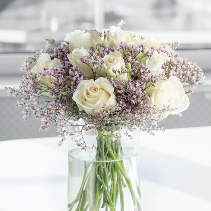 This image captures an elegant sympathy bouquet that seems to breathe calm into the room, composed primarily of creamy white roses and soft lavender-toned filler flowers. About a dozen roses are in full bloom, their petals open wide yet still cupped in classic, spiralling formations that draw the eye gently towards the centre of each flower. The colour is a warm off-white, leaning delicately towards ivory, evoking purity without feeling stark. Between the roses, airy tufts of tiny pastel blossoms create a cloud-like surround; their hues shift subtly from pale lilac to muted pink, and their smaller, papery petals bring texture and volume to the design. All the blooms emerge from a tall, slim cylindrical vase made of clear glass, its surface catching the light and echoing the clean lines of contemporary flats around Clerkenwell and Farringdon. Through the glass, the arrangement's structure is laid bare: neat, green stems cut cleanly at the base and fanning out gracefully into the clear, shallow-gleaming water. The bouquet stands on a bright, uncluttered white surface that reflects soft light back into the arrangement, while the background fades into a gentle blur of whites and pale greys, suggesting a quiet indoor setting-perhaps a reception room near Clerkenwell Green or a small studio overlooking St John's Square. The mood is subdued and reflective, conveying sympathy and remembrance without excess. This is the kind of thoughtful piece often chosen to express condolences, to sit beside a framed photograph, or to offer a subtle yet heartfelt gesture of support in times of loss across the Clerkenwell community.