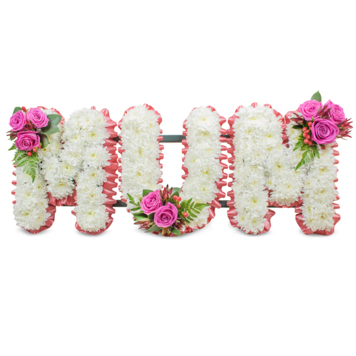 A thoughtfully made sympathy tribute displaying the word MUM in three connected capital letters, photographed on a plain white surface that highlights the arrangement's neat craftsmanship. The main bodies of the letters are built from tightly packed, immaculate white chrysanthemums whose small blooms nestle together to form a pillowy, consistent texture across each letterface. Along each outer edge a soft, ruffled pale-pink ribbon or cloth trim frames the letters, introducing a gentle halo of colour that complements the white without overwhelming it. A faint dark green support armature is visible between the letters, suggesting the standing construction used for memorial displays. The arrangement is enlivened by three coordinated accent posies: deep fuchsia-pink roses - some opening, some in bud - are placed at the upper left of the initial M, at the centre curve of the U, and on the upper right of the final M, each grouping accompanied by glossy green foliage (fern-like fronds) and clusters of small, coral-red hypericum berries which add sheen and a subtle tactile counterpoint. The overall impression is one of respectful tenderness: the cool, clean whites convey solemnity while the pink roses and ribbon bring warmth and affection. I picture this piece standing near a memorial table at a Clerkenwell funeral, perhaps within sight of the Barbican, where its restrained scent of chrysanthemums and roses and its balanced silhouette would offer comfort and an eloquent expression of mourning and love.