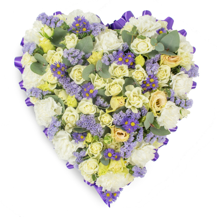 Lovingly composed on a white backdrop, this heart-shaped memorial wreath combines florist's precision with an intimate, consoling mood - exactly the kind of tribute we prepare at Clerkenwell Flowers for services around Clerkenwell Green and Farringdon. The dense center is woven from cream and pale yellow roses, their petals ranging from tight, honeyed buds to softly opening blossoms that add depth and a velvety tactile quality. Intermingled white carnations bring generous, cushiony volume and a timeless sympathy tone. Lacy clusters of lavender statice drift through the arrangement, their small, papery flowers introducing a gentle, cool violet texture, while tiny purple daisy-like asters with bright yellow eyes create cheerful, contrasting points of colour that prevent the palette from feeling one-dimensional. Blue-green eucalyptus leaves are threaded throughout; their rounded, silvery foliage punctuates the massed blooms and suggests a subtle herbal freshness when held close. Around the edge, a continuous, looped purple satin ribbon has been carefully folded and secured to form an elegant frame, the ribbon's lustre lending formality and a finished silhouette. The overall impression is serene and respectful: a tactile, fragrant composition made to convey enduring love and remembrance. Suitable for funerals, memorial services or graveside placement, this heart-shaped tribute offers a measured, heartfelt way to honour someone's memory in and around Clerkenwell's local churches and chapels.