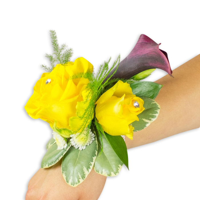 Presented on a light-to-medium forearm and shot against a bright white backdrop, this elegant wrist corsage combines sunny yellow roses with a single, dramatic deep-purple calla to striking effect. Two yellow roses anchor the arrangement - the left bloom fuller and more open, the right one slightly smaller and more cup-shaped - each petal soft and layered, the surface catching a gentle wash of studio light. Near the centre of each rose sits a tiny clear rhinestone, a polished accent that glints with movement and underlines the corsage's formal intent. The calla lily, almost black-cherry in hue, lifts behind the roses and curves delicately upward and to the right, its velvety inner surface contrasting with the roses' softness. Surrounding greenery adds contrast and structure: feathery asparagus fern threads through the top and back of the composition, adding a light, airy texture, while broader variegated leaves with pale margins create a supportive base that reads as cool and glossy. Small white button-like filler flowers are tucked in to increase warmth and fullness, softening the transitions between focal blooms and foliage. The band fastening the arrangement is slim and mostly hidden beneath the flowers, presenting the corsage as an integrated adornment worn diagonally from the lower-left toward the centre-right of the image. With a crisp, accessible presentation, this design evokes quiet celebration - perfect for prom nights, wedding parties, or a ceremony in or around Clerkenwell Green - and highlights the tactile contrasts between velvety petals, satin calla, and feathery green accents.
