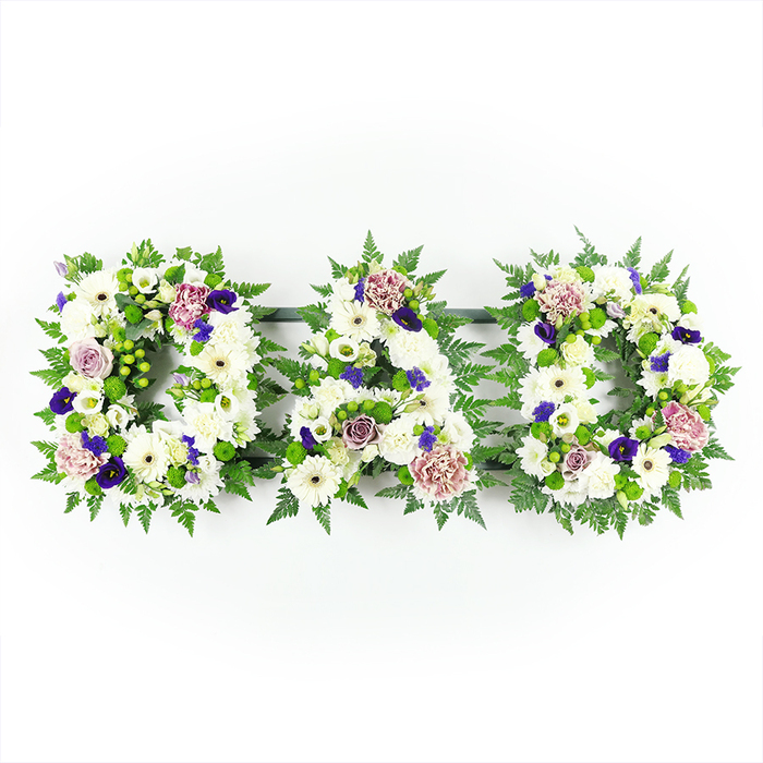 The image shows a traditional yet beautifully detailed floral lettering tribute spelling out DAD, created as three separate but perfectly aligned capital letters set horizontally on a stark white background. Each letter is packed so tightly with flowers that no mechanics show through: white chrysanthemums form the main body, their full, generous blooms creating a smooth and comforting surface, while white gerbera daisies add a slightly larger, daisy-like face with soft greenish centres. Interspersed among these are delicate white lisianthus and short-stemmed spray roses, adding variation in petal shape and a touch of refined elegance. Framing the letters is a collar of lush green foliage, largely made up of feather-light fern fronds and structured leafy stems that hold the outline in a crisp, readable form. Dotted within the green are bright lime-green button chrysanthemums and berry-like accents, lending a fresh, almost springlike feeling despite the solemn purpose. Deeper purple tones run like a quiet thread through the design: small velvety blossoms reminiscent of pansies, hints of purple spray carnations, and soft lavender-mauve roses that introduce warmth and emotional depth. The lighting is even and clear, similar to natural daylight filtering into a Clerkenwell studio near Farringdon, revealing the intricate textures without strong shadows. This carefully composed DAD tribute feels deeply personal, suited to rest at the front of a funeral service, to travel with the hearse through local Clerkenwell streets, or to be placed at a graveside as a lasting symbol of love, gratitude, and remembrance for a cherished father.