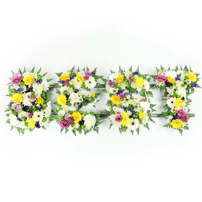 A carefully crafted floral tribute spells out the word BEST in bold, garden-rich letters set against a pure white background, photographed directly so every textural nuance is visible. The palette is immediately comforting: crisp whites and golden yellows dominate, punctuated by gentle lavenders and bright, magenta-pink notes. Large, cushiony white chrysanthemum-like flowers and creamy gerberas make up the soft, luminous foundation of each letter, while vibrant yellow roses and gerbera daisies lift the arrangement with sunny clarity. Deeper purple lisianthus or small chrysanthemums provide a sober, elegant depth, and sprays of pink roses or carnations offer cheerful, affectionate highlights. Small green and white filler clusters create delicate transitions and tactile interest across the densely packed surfaces. Dark green fern fronds are generously worked around and between letters, their fine blades creating a soft, leafy frame that contrasts with the full, rounded petals. Through small gaps the dark green or subtle metal structural bars are visible, particularly along the E and the T, a reminder of the skill that secures such large-scale floral lettering. The lighting is bright and even, accentuating the freshness and crisp contours of each bloom and giving the piece an air of calm dignity. This composition reads as a sympathy or memorial tribute - a tender way to honour a loved one - yet its uplifting yellows and vibrant pinks can also translate to celebration. I imagine it delivered with care across Clerkenwell, perhaps placed beside a service at Spa Fields or sent to a family home near Farringdon Road.