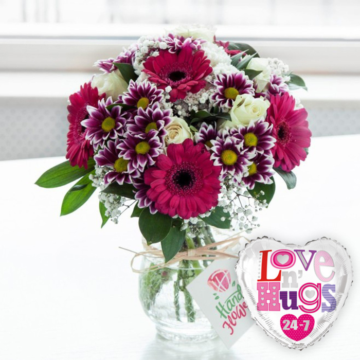 A bright, celebratory flower display is arranged on a pale surface that could easily be a kitchen table or desk in a Clerkenwell apartment flooded with natural light. At the centre, large gerbera daisies in vivid magenta and fuchsia radiate outward, their wide petals smooth and velvety, surrounding dark, almost chocolate-coloured centres that add sophistication to the otherwise playful palette. Around these, tight clusters of decorative chrysanthemums create intricate rings of colour: deep purple toward the middle, fading neatly to white at each petal tip, with warm yellow centres that pop like tiny suns. Creamy white roses are threaded throughout the design, some in tight buds and others opening into soft spirals, giving the bouquet an air of classic romance. Sprigs of white gypsophila, or baby's breath, drift through the arrangement in tiny clouds, breaking up the stronger hues and lending a delicate, lace-like texture that softens the overall shape. Lush green foliage, glossy and fresh, frames the blooms and emphasises the rounded, generously full form of the bouquet, the kind a local florist might prepare at Clerkenwell Flowers for a birthday or 'thinking of you' delivery. The stems are gathered neatly into a clear, stout glass vase filled with clean water, the cut ends visible and orderly. A piece of rustic, natural-coloured twine is tied around the neck of the vase, knotted in a simple bow and holding a small, rectangular white tag. On the tag, a stylised pink rose icon rests above friendly green script, evoking something hand-written and personal. Leaning just in front and to the right, a polished silver heart-shaped foil balloon catches the light, its white centre printed with the phrase Love n' Hugs 24-7 in lively blocks of patterned pink, purple, and blue letters, small heart motifs tucked among them. The softly blurred background suggests a bright window looking out toward Clerkenwell's streets, creating a warm, affectionate atmosphere perfect for romantic surprises, get-well wishes, or everyday gestures of care.