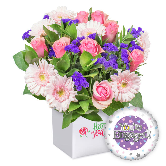Bright due to foreground light, a vivid engagement bouquet stands tall in a snowy white square box that frames the petals like a gallery piece. The pink roses are a chorus of romance, from tight buds to ripe blossoms, paired with light pink gerberas edged in white and with pale yellow centers. Purple filler blooms provide dramatic pops, while generous green foliage adds depth and a garden feel. The arrangement rests in a clean box with a discreet red rose emblem on the front and green script, a nod to the craft of a Clerkenwell florist without naming it. To the lower right, a shiny round foil balloon completes the set, its central purple disk bearing the silver inscription You're Engaged!, with the golden O forming a ring to celebrate the proposal. The white outer ring is speckled with pastel hearts and small stars, echoing the festive mood.
This is a complete engagement gift: a bouquet that works beautifully on a table at a flat near Smithfield Market or in reception at a Clerkenwell coworking space. The colours blend softly yet jubilantly, making any room feel brighter and more hopeful. The piece has natural movement-the stems align in a precise, yet loose arrangement-testifying to the skill of the maker and the care that's gone into choosing each bloom for freshness and longevity, with delivery available across Clerkenwell and the surrounding boroughs.