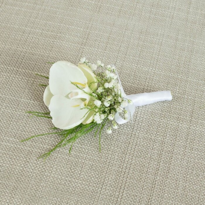 An elegant white orchid boutonniere is displayed on a pale beige, woven fabric surface, the subtle texture of the cloth providing a calm, natural backdrop similar to the textiles used in Clerkenwell's creative studios. At the centre of the piece is a flawless Phalaenopsis orchid bloom, its petals broad, gently curved, and slightly glossy, catching the light with a soft sheen. The orchid's throat reveals a delicate touch of buttery yellow and a hint of cream, lending warmth and dimension to the otherwise crisp white flower. Surrounding the main bloom is a halo of tiny baby's breath blossoms, each minuscule white flower forming part of an airy, cloud-like collar that softens the silhouette and adds a sense of romance and lightness. Threaded through this halo are slim strands of fresh greenery, likely a fine fern or similarly delicate foliage, their vivid green tones creating a graceful contrast against the white petals and pale background. The stems are gathered tightly and wrapped with care in a smooth, matte white satin ribbon, the wrapping meticulously even and finished with a discreet, tidy bow that suggests skilled hands at work, perhaps in a local workshop just off Farringdon Road. The lighting in the image is gentle and diffused, with no harsh highlights, allowing the textures of the orchid, the baby's breath, and the ribbon to be clearly seen and appreciated. The boutonniere feels perfectly poised for a wedding or formal celebration in Clerkenwell, from chic city ceremonies to intimate gatherings, embodying a sense of timeless sophistication, purity, and thoughtfully curated design for a groom, best man, or honoured guest.