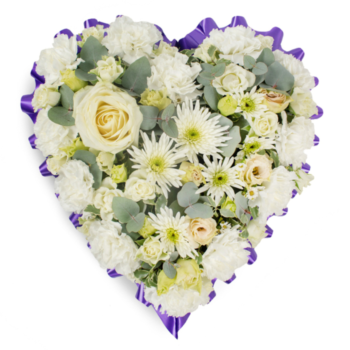 A refined heart-shaped funeral arrangement occupies the frame, its dense surface a study in white and cream textures and soft contrasts. The centre is anchored by one creamy-white rose, its compact, velvety petals spiralling inward like a private gesture; radiating from that focal bloom, clusters of white carnations create a voluminous, cloud-like body, their ruffled forms marrying softness with structure. Interspersed are white chrysanthemums - some with long, narrow 'spider' petals and subtle lime-green eyes - which introduce an energetic, almost architectural detail against the smoother roses and carnations. Smaller cream roses and delicate lisianthus buds nestle in the crevices, lending intimate scale and the suggestion of unfolding memory, while blue-green eucalyptus leaves are strategically placed to give cool tone, depth and a faint herbal fragrance. The heart's silhouette is crisply defined by a deep purple ruffled ribbon, its scalloped edge providing a formal, respectful border that both contains and elevates the pale blooms. The arrangement is captured in gentle, even light that reveals petal texture, shadowed recesses and the careful hand-placing of each element; it feels tactile and quiet, a composition meant to be felt as much as seen. Commonly chosen for memorial services around Clerkenwell - from Spa Fields to the churches on St John Street - this piece conveys solemnity, tenderness and a timeless expression of condolence, a floral elegy that honours memory with grace.