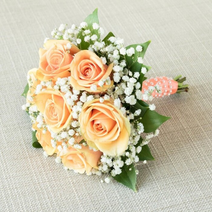 Showcased against the inviting backdrop of oatmeal-hued linen often found in the homes and studios of Clerkenwell, this hand-tied bouquet radiates warmth and quiet luxury-an ideal companion for bridesmaids or the romantic at heart. At its core lie seven to eight roses, each a soft masterpiece of pale apricot and luminous peach, their petals tightly spiraled in the centre and gradually blossoming outward in layers of gentle elegance. Between these radiant blooms, generous sprays of white baby's breath (Gypsophila) drift like clouds, offering a diaphanous contrast to the roses' warmth and evoking the airy, hopeful feeling of Clerkenwell mornings. The bouquet is given depth by the presence of deep green rose leaves, glossy and fresh, offering a foundation of calm amidst the soft riot of colour. Stems are neatly trimmed and tightly bound with a coral-peach satin ribbon, its surface adorned by a delicate spiral of lustrous white pearls-a tactile and visual flourish, reminiscent of the artisanal detail found in the neighbourhood's hidden boutiques. The rounded shape of the bouquet, expertly constructed, rests horizontally and slightly angled to catch the gentle, diffused light, which flatters the subtle tonal shifts and textures within the arrangement. This design, ideal for a wedding on Amwell Street or a celebration in Charterhouse Square, is lovingly composed by Clerkenwell Flowers, ready to lend its charm, sophistication, and romance to special moments across the area.