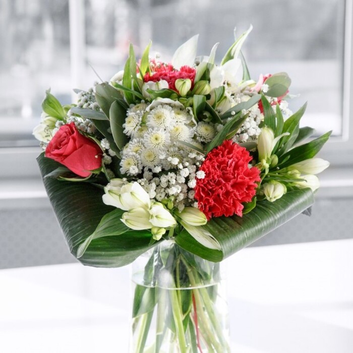 In this image, a sophisticated red and white bouquet stands in a clear cylindrical glass vase, the water line crisp and transparent, revealing straight, freshly trimmed green stems arranged with deliberate care. Rising above the vase, the flowers form a rounded, abundant canopy of colour and texture. Deep red roses, with their velvety petals unfolding in tight spirals, anchor the composition, their rich tone suggesting love and intensity. At the front, a generously layered crimson carnation, all ruffles and fine edges, creates a focal point that draws the eye immediately. Interspersed between these bold, romantic reds are cool, calming whites: slender-petalled alstroemeria patterned with subtle speckles, rounded white blooms reminiscent of small chrysanthemums, and several firm, unopened white lily buds that suggest the bouquet will evolve over the coming days. Throughout, clouds of white gypsophila drift like fine lace, softening the contrasts and adding an airy lightness. The arrangement is framed and supported by lush foliage. Broad, glossy dark green leaves are carefully wrapped and arched at the base to form an elegant collar, while eucalyptus-like sprigs and pointed green leaves introduce varying heights, shapes, and shades of green, giving the bouquet depth and movement. The flowers rest on a smooth white surface, bathed in gentle, diffused daylight similar to the soft light that filters into Clerkenwell homes overlooking Spa Fields or Hatton Garden. The background is bright and out of focus, ensuring the bouquet remains the clear star of the scene. The mood is one of quiet yet confident celebration, ideal for a dinner table in a Clerkenwell loft, a thoughtful anniversary gift, or a gesture of heartfelt thanks to someone special nearby.