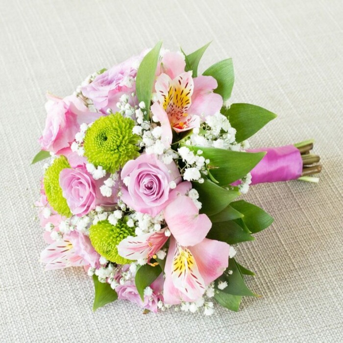 In this radiant, close-up image, a hand-tied bridal bouquet sits diagonally on finely woven beige linen, its elegant details revealing the artistry of local Clerkenwell florists. Soft pastel pink roses, some just opening, others in luxuriant full bloom, fill the arrangement's center, their velvety spirals inviting a lingering gaze and evoking the classic romance of garden gatherings in St John's Square. Vivid lime-green button chrysanthemums add a bold, textural accent, their tight, round forms playfully contrasting the softness of the roses. Light pink alstroemeria, with gentle streaks of yellow and delicate maroon speckles, open widely amidst the ensemble, their exotic look uplifted by the bouquet's modern palette. Tiny clouds of baby's breath (gypsophila) are sprinkled throughout, infusing the arrangement with airiness and tying together the pink, green, and white for a harmoniously balanced look. Deep green leaves are woven carefully among the blossoms, providing both structure and a lively frame that hints at the local greenery of nearby parks. The bouquet's natural stems are expertly gathered and bound with a lustrous magenta satin ribbon, knotted gracefully and leaving the clean-cut ends visible for a contemporary yet organic effect. The lighting, soft and ambient, highlights each petal's freshness and the interwoven textures of flower, foliage, and ribbon. This bouquet speaks to Clerkenwell's sophisticated blend of history and innovation-inspired by its vibrant community and perfect for a spring wedding, thoughtful anniversary, or celebratory gift.