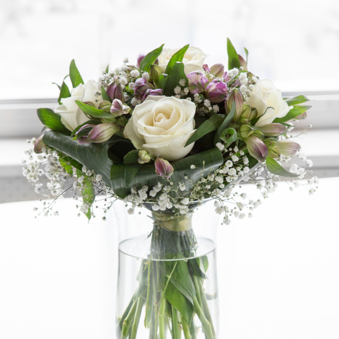A graceful, understated bouquet of white and purple flowers stands in a clear glass vase, illuminated by soft, bright light that smooths away any harsh shadows and lends the entire scene a quiet, respectful calm. At the centre, creamy white roses unfurl their petals in perfect spirals, their surfaces smooth and almost luminous, suggesting innocence and enduring affection. Tucked between these focal blooms are alstroemeria in shades of lavender and muted violet, some petals edged with green, their slender shapes adding texture and subtle movement to the design. Feathery sprays of white baby's breath weave in and out among the larger flowers, filling small spaces and creating a gauzy, cloud-like effect that makes the bouquet feel light and comforting rather than heavy or overdone. Encircling the blooms is a generous collar of deep green foliage, a mixture of glossy broad leaves and more delicate stems that rise and curve gently, framing the pastel tones in a natural, supportive embrace. Through the cylindrical vase, the straight green stems are visible, resting in clear water that hints at the freshness and care behind the arrangement, much like a bouquet prepared that morning in a Clerkenwell workshop before being sent along Rosebery Avenue or towards Farringdon. The background is a softly diffused white, suggesting a well-lit room or studio, which allows the colours and textures of the flowers to stand out without distraction. The mood is tranquil and composed, making this arrangement fitting for sympathy, remembrance services, or as a gentle presence in a quiet room where comfort and subtle beauty are needed most.