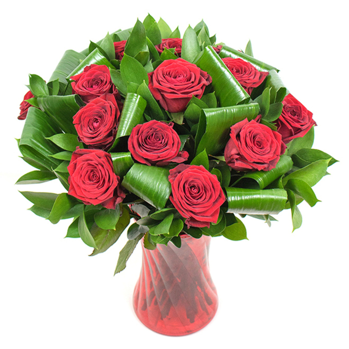 This carefully composed arrangement presents approximately a dozen deep crimson roses, each bloom opened to reveal tightly spiraled, velvety petals that absorb and reflect light like plush fabric. The roses form a lush, rounded mass, their rich red tones intensified by soft, even illumination that brings out petal texture and subtle tonal variation from core to edge. Surrounding the blooms is an abundance of dark green foliage: smaller, glossy leaves create a verdant cushion while broader leaves - reminiscent of Aspidistra - are expertly rolled and curved into sculptural cylinders and loops. Those shaped leaves are not merely filler but structural elements, artfully placed to cradle stems, add height, and introduce elegant architectural movement that modernises the classic red-rose silhouette. A tall, translucent ruby-red glass vase with a gentle waist and a faint ribbed twist completes the composition; its vertical lines echo the stems and reflect light in a way that unifies vase and flowers. The whole piece is photographed against a pure white background so the deep reds and emerald greens read with vivid clarity. Crafted in our Clerkenwell studio by experienced hands at Clerkenwell Flowers, this bouquet balances traditional romance with contemporary design - ideal for anniversaries, Valentine's gestures, or refined venue styling near Charterhouse Square or Smithfield. The image invites you to imagine the soft, heady scent of rose and the smooth, cool feel of glass beneath your fingertips, promising a memorable floral moment for someone special.