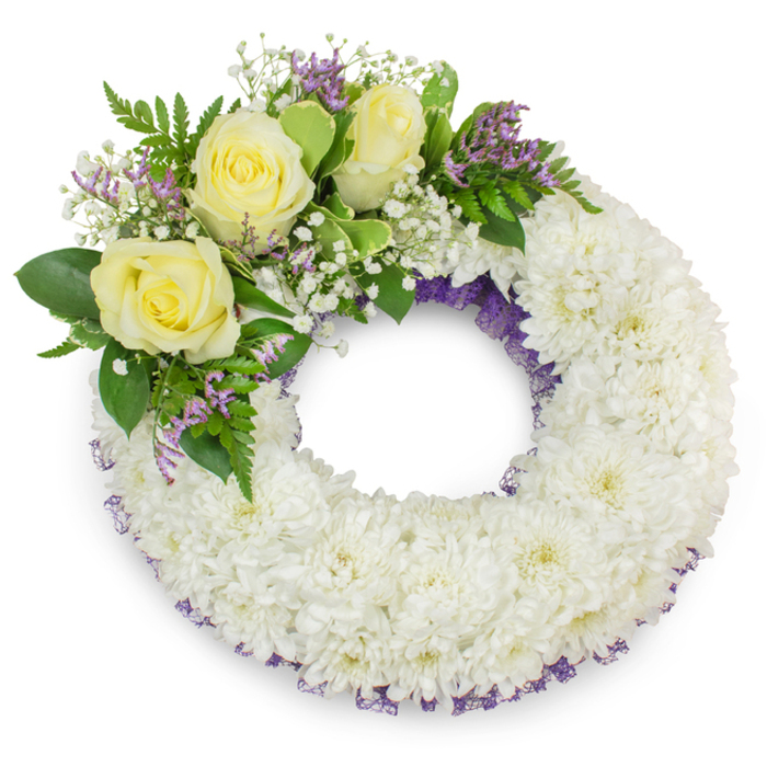 A dignified, circular funeral wreath sits against a pure white studio backdrop, its form defined by hundreds of small, pristine white spray chrysanthemums tightly set to produce a uniform, velvety field of petals that conveys calm and reverence. The wreath keeps a perfectly open centre and carries a faint, elegant border of dark purple netting or ribbon that shows through the chrysanthemum heads along both the inside and outside edges, giving subtle structure and depth. Anchoring the design at the upper left is a tasteful cluster of three creamy yellow roses, each bloom layered and soft-edged, symbolising care and remembrance; these roses are complemented by misty sprigs of white gypsophila which add a cloud-like lift and by delicate clusters of lavender-toned statice or limonium that offer a restrained, slightly textural colour note. A mix of foliage - broad glossy leaves juxtaposed with finer, ferny fronds - frames the accent bouquet, adding cool green tones and a natural, leafy scent. The lighting is even and soft, producing a gentle shadow beneath the wreath that gives it presence on the white plane and reveals the careful mechanics of design: graduated blossom sizes, hidden wiring, and considered balance so the wreath sits level and graceful. This piece reads as an eloquent tribute for a funeral, wake, or memorial, suitable for Clerkenwell families requesting delivery to local venues such as the Union Chapel or a nearby cemetery; it speaks of peaceful remembrance and tactful floral restraint.