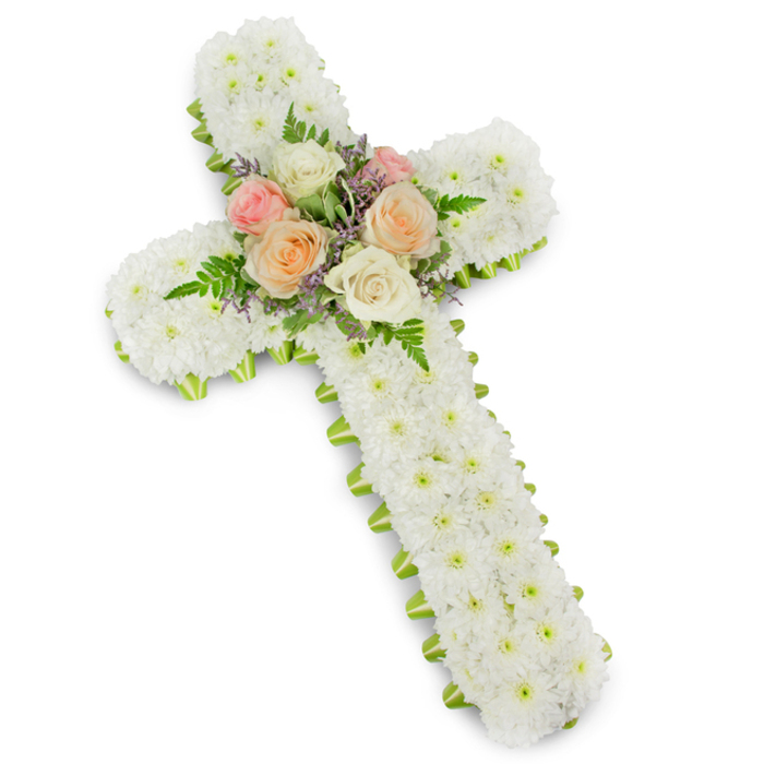 The image presents an elegant funeral flower cross, photographed against a clean white backdrop that subtly echoes the clarity and stillness of a quiet morning in Clerkenwell. The cross itself is completely covered with white button chrysanthemums, each tiny bloom placed closely beside the next so the surface appears dense and cushiony, with no visible gaps. This blanket of white suggests purity and peace, the rounded petals catching the soft light to create gentle shadows within the floral mass. Running along the very edge of the cross is a slim band of lime green ribbon, carefully wrapped so that only a sliver of colour outlines the shape. This fresh green detail frames the design much like the borders of a stained-glass window in a local church, giving structure without overshadowing the purity of the white flowers. At the intersection of the vertical and horizontal arms sits a carefully crafted cluster of roses, forming the emotional centre of the arrangement. These roses are open and velvety, displaying shades of pale blush pink, warm peach, and creamy ivory that transition smoothly from one tone to the next. They are nestled among light, feathery fern fronds, whose fine texture breaks up the expanse of chrysanthemums, and threaded with tiny sprays of lavender or soft purple filler flowers. The composition feels balanced and gracious, clearly designed as a sympathy tribute for funerals, memorials, or graveside services. It suggests a message of faith and eternal rest, an arrangement one might choose when sending condolences or arranging a final farewell for a loved one from the Clerkenwell community.