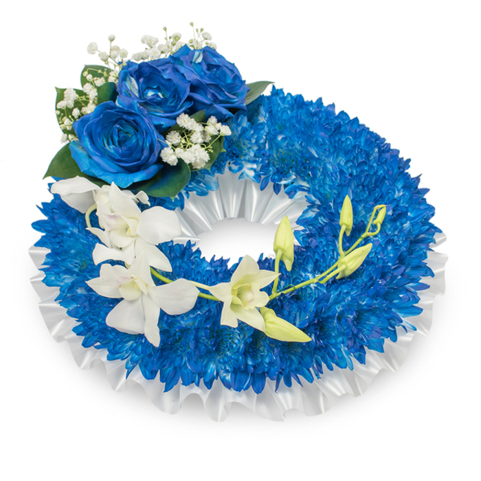 An elegant circular wreath presented against a pristine white backdrop, carefully composed to convey respectful remembrance. The dominant surface is a thick ring of intense royal blue chrysanthemums, each tiny, radiating petal contributing to a voluminous textured base that feels both composed and consoling. Inside the wreath, a ruffled white satin ribbon is pleated along the opening, its soft sheen and modest folds lending a classical framing to the saturated blue. At the upper-left curve a trio of large blue roses forms a quiet focal point; their lush, velvety petals unfold in a deep sapphire that harmonises with the chrysanthemums. Delicate sprays of white gypsophila and a few glossy dark green leaves tuck around the roses, adding air and a traditional sympathy accent. Descending from mid-left toward the lower right, a string of ivory-white orchids drifts across the wreath, their broad, graceful petals and columned centres offering a serene counterpoint to the dense blue. Interspersed among the orchids are bright green buds in differing stages of bloom, small hints of life and continuity threaded through the composition. The feeling is hushed and composed, the cool blues calling to peace and memory while the whites speak to purity and solace. The textures - spiky and plush, velvety and satiny - are handled with careful craftsmanship you would expect from a local Clerkenwell florist; Clerkenwell Flowers often places similar tributes for services around Smithfield and the Barbican, where families seek a dignified expression of condolence.