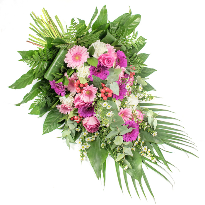 Against a clean white studio background, a lush, asymmetrical funeral spray stretches diagonally from the upper left to the lower right, creating a sense of gentle movement and flow. The composition is rich but controlled, carefully balancing vibrant colour with serene whites and abundant greenery. Large gerbera daisies in soft blush pink and strong fuchsia form the visual anchors of the piece, their round, open faces catching the light and bringing a quiet optimism to the tribute. Around them, elegant pink roses with layered, spiralling petals nestle into the greenery, their more traditional form adding a timeless note of remembrance. Clusters of ruffled white blooms, reminiscent of mini carnations or dianthus, scatter through the arrangement like pockets of brightness, while sprigs of tiny white daisy-like flowers meander between stems, creating a delicate, almost meadow-like feel.
Threaded among the blooms are bright red hypericum berries, their smooth, bead-like forms providing subtle bursts of colour and a touch of natural texture. The flowers are supported by a generous framework of foliage, echoing the green pockets found around Clerkenwell's gardens and courtyards: broad, glossy leaves form a strong base; airy fern fronds add softness and a feathery edge; and rounded, silvery eucalyptus leaves introduce a cool, contemporary accent and suggest a refreshing scent. At the lower right, long palm fronds sweep outward, enhancing the spray's elongated, cascading profile, while at the upper left, the neatly banded stems show the craftsmanship behind the hand-tied design. This refined funeral spray, suitable as a casket decoration or a significant standing tribute at local Clerkenwell services, conveys deep sympathy and respect, offering a colourful yet dignified way to honour a loved one's life.
