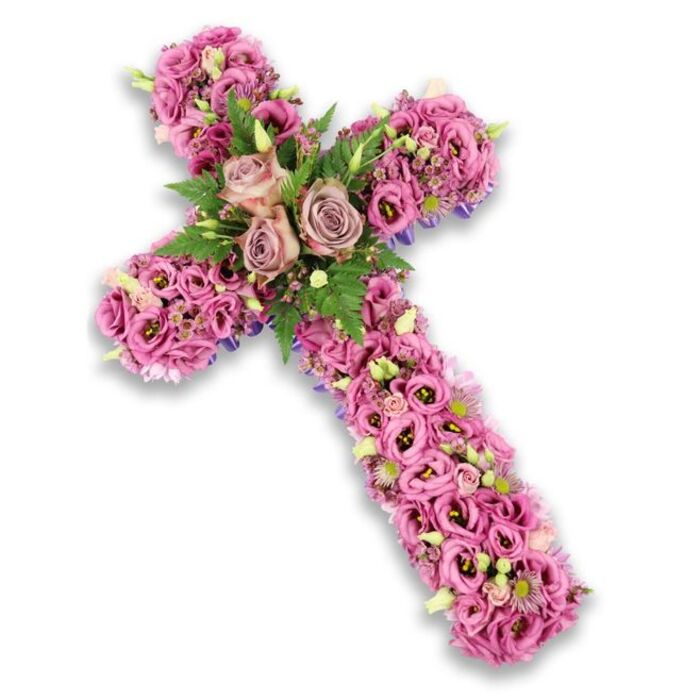 A meticulously crafted floral cross tribute is displayed on a stark, bright white background, its entire surface transformed into a tapestry of soft pink, mauve, and lilac blossoms that convey both tenderness and reverence. The cross itself is three-dimensional and generously proportioned, each arm and the full length densely packed with lisianthus flowers whose ruffled petals interlock, creating a cushioned, almost cloud-like texture. The colours drift from the palest blush and gentle lavender to deeper rose-pink, with some blooms showing darker centres that introduce quiet contrast and a sense of depth. At the exact point where the vertical and horizontal arms meet, three prominent roses in a sophisticated dusty rose or muted mauve shade form the emotional centre of the design. Their fully unfurled petals spiral outward, suggesting openness and enduring love, while being softly cradled by fern fronds and small, vivid green leaves that bring the arrangement to life. Interspersed throughout the cross are tiny clusters of filler flowers in pinks and purples, along with light green buds and a scattering of petite white blossoms, each adding delicate texture and interest when the eye travels across the piece. Near the edges, particularly along the outer contours of the cross, slender strips of darker purple ribbon or fabric peek out from below the blossoms, subtly outlining the sacred shape and adding a refined finish. The simple, shadowed white background allows the colours and craftsmanship to stand out, as if the piece had just been prepared in a calm studio off St John Street. This floral cross, the kind of tribute a Clerkenwell family might choose for a funeral or graveside service, speaks quietly of faith, remembrance, and a deep, enduring respect for the one who has passed.
