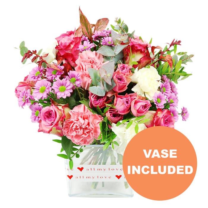 A richly detailed floral arrangement fills the frame, set sharply against a clean white background that makes every colour and petal shape stand out, much like a display in a Clerkenwell design studio. The bouquet is arranged in a modern, square glass vase with softly rounded edges, its base dressed in a white band printed with repeating bright red hearts and the sweet phrase all my love in flowing script. Inside the vase, the stems are visible, cleanly cut and carefully layered to support the abundant display above. The main impression is one of pink richness: large roses in deep fuchsia and softer blush pink rise proudly from the centre, some fully open to reveal intricate, velvety petals, others still cupped in bud form, suggesting freshness and longevity. Surrounding them, ruffled carnations in tones of coral, salmon, and peach-pink add soft volume, their many frilled petals catching the light and creating delicate shadows. Interspersed between the larger blooms are smaller, daisy-like chrysanthemums in shades of lavender and purple, each with a vivid lime-green centre that introduces a lively, almost sparkling contrast. Creamy white carnations and petite white daisies provide breathing space between the stronger colours, lending an elegant harmony to the overall palette. The flowers are cradled by generous greenery: rounded eucalyptus leaves with a silvery-green hue, deeper green foliage for structure, and a few reddish-brown leaves that add a touch of depth, reminiscent of autumn walks near Clerkenwell Close. The bouquet forms a balanced, rounded silhouette, neither too tall nor too wide, making it ideal for a dining or side table. In the bottom right corner, an orange circular graphic clearly reads VASE INCLUDED in bold white capital letters, signalling that this is a complete, ready-to-display floral gift suitable for delivery to homes, studios, and offices throughout Clerkenwell.