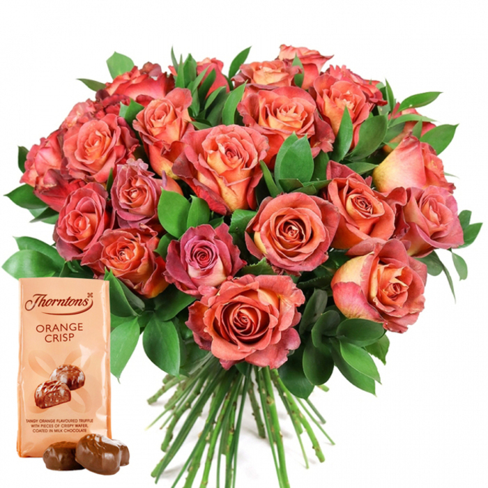 Expertly arranged against a pristine white setting, this generous gift set features a sumptuous rose bouquet and a pouch of Thorntons Orange Crisp, thoughtfully curated by Clerkenwell's experienced florists. The bouquet forms the visual centrepiece, brimming with dozens of freshly cut roses in an enlivening palette that drifts from creamy peach in the heart of each bloom to the saturated coral and rosy-pink edges along the flared outer petals. Subtle bicolor transitions and ombre blending lend a sense of artistry akin to the dynamic murals found on Clerkenwell Road. Nestled amidst velvety, dark green foliage-likely ruscus or local salal chosen for sheen and longevity-the roses seem to glow with inner light, their arrangement dense yet balanced. The pouch of Thorntons Orange Crisp occupies the lower-left, its pale beige design marked with an elegant logo and a tempting depiction of smooth chocolate truffles, described as orange-flavored and wafer-studded within a silky coating. In front, two freshly unwrapped chocolates sit on the surface, their rounded, glossy forms hinting at decadent pleasure. The bouquet's neatly gathered, extended green stems speak to professional care, suggesting freshness ideal for same-day delivery across Clerkenwell and Farringdon. This image invites feelings of warmth and thoughtfulness-perfect for celebrating milestones, sending appreciation, or expressing love within London's historic creative quarter.