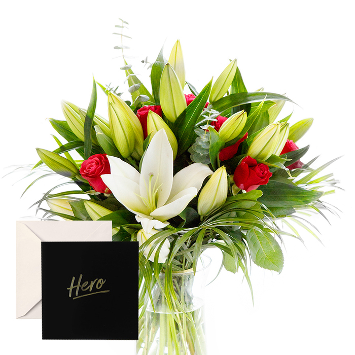 A bold, uplifting arrangement that pairs the calm of white lilies with the fierce warmth of red roses, designed to toast achievement and resilience. The white lilies lie near the center, their luminous petals like soft arctic light, while the red roses rise like embers among abundant deep green foliage. The surrounding leaves create a forest-like backdrop that grounds the bouquet with a cool, verdant energy. Unopened buds add anticipation, inviting the eye to follow the stems upward. Light falls across the blooms, highlighting crisp contrasts and the decadent textures of petals and silk-like leaves. It feels crafted for a moment of triumph, perhaps a milestone reached near Smithfield Market or a major project celebrated at a Clerkenwell studio, yet appropriate for a personal thank-you, a grant of recognition, or a festive send-off. The bouquet is arranged in a clear vase that emphasizes the stems' height variation, a hallmark of the hand-tied technique. The card marked 'Hero' tucked into the arrangement seals the message: you inspired courage and care in others. Available for same-day delivery across Clerkenwell, Holborn, and surrounding wards, this gift makes a vivid, confident statement-special, memorable, and distinctly local.