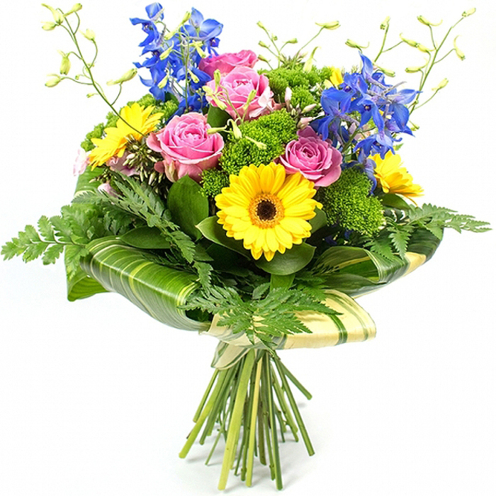 A fresh, professionally arranged bouquet stands upright against a seamless white background, its colours clean and vivid, as though just prepared in a Clerkenwell flower shop before being sent out for local delivery. Dominating the centre are several full, pink roses, their petals layered in gentle spirals that suggest softness and affection, each bloom slightly different, adding a natural, unforced charm. Encircling and intermingling with them are bright yellow gerbera daisies, each flower head a tidy disc of slender petals radiating from a dark brown core, like small suns beaming warmth into the arrangement. Rising from within and just behind the central cluster, tall stems of deep blue delphinium carry multiple petite florets along each spike, introducing vertical structure and a cool, inky contrast reminiscent of evening skies over nearby Clerkenwell Green.
Around this colourful heart, an assortment of greenery creates depth and balance. Long, feathery fern fronds trail gently from the outer edges, softening the silhouette and adding a slightly wild, garden-inspired feel. Compact clusters of bright green, cushiony filler flowers are tucked between the roses and gerberas, building volume and textural interest without overpowering the main blooms. At the lower portion, broad, glossy leaves-some subtly striped in lighter shades of green-are wrapped artfully around the base of the flower heads, forming a neat, architectural collar that feels modern yet organic. Above the main blooms, a few slender green stems stretch upward, tipped with tiny unopened buds that hint at freshness and new beginnings. The natural stems are visible below, tightly gathered and bound in a pale cream or soft green wrapping material, secured with a simple bow. The overall effect is that of a joyful, contemporary bouquet, perfectly suited to brighten a Clerkenwell apartment, celebrate a work promotion near Farringdon, or bring a splash of colour to a dinner table overlooking St John's Square.