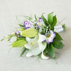 An elegant, gently elongated bouquet is laid diagonally across a pale beige, textured cloth, as if waiting to be lifted for a wedding procession through Clerkenwell's cobbled streets. The composition is dominated by large white lilies, their petals smooth and softly luminous, opening wide to expose warm yellow-orange stamens and delicate freckles near the throat. Nestled around them are vivid green lily buds, still closed but full of promise, adding a sense of anticipation to the scene. Between these focal blooms, Lisianthus flowers weave in and out, their petals ruffled and cup-shaped, white at the base and edged in vibrant purple that ranges from fresh lavender to rich violet, giving the bouquet a modern, artistic flair. Scattered throughout are airy clusters of baby's breath, each miniature white flower contributing to a cloud-like halo that softens the structure. The greenery is lush and layered: broad, dark green leaves provide depth and form, while fine, trailing fronds and a few bolder, spiky stems lend movement and a slightly untamed grace, echoing the blend of old and new architecture around Clerkenwell Green. The gathered stems are wrapped in smooth white satin ribbon, delicately accented with tiny pearlescent beads that suggest a bridal or ceremonial purpose rather than a casual posy. Soft, even light bathes the arrangement, highlighting the fresh textures and crisp colour contrasts against the calm, neutral backdrop. The overall impression is of refined but approachable beauty, the sort of bouquet one might carry down the aisle at a local ceremony or order for same-day delivery around Farringdon and St John's Street to celebrate love, commitment, or a quietly joyful milestone.
