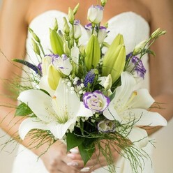 In the gentle light of a Clerkenwell morning, a bride's bouquet prepared for a ceremony on Exmouth Market glows in delicate harmony between tradition and innovation. Creamy Oriental lilies, bold and fragrant, open fully-revealing gently speckled petals with sunlit yellow stamens, each blossom a gesture of devotion. Around them dance lisianthus flowers, some sugared white with feathery, lavender-tipped edges, others still curled in tight green buds with a promise of future bloom. Wisps of pure white gypsophila create clouds of softness between the deeper, textural spikes of violet-hued veronica, all resting among fresh green foliage and graceful asparagus fern. The bouquet, gathered with understated care by Clerkenwell's local florists, nestles securely in the bride's soft, lightly polished hands, her anticipation echoing in the layers of blooms. The surrounding blur of her white, strapless dress frames the arrangement, allowing each petal's subtle glow to be appreciated. With artistry and care, this wedding bouquet celebrates both the vibrancy of local London life and the quiet joy of union, ideal for couples seeking a refined yet heartfelt flourish from Clerkenwell to Bloomsbury Square.
