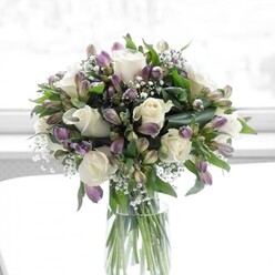 A graceful bouquet of creamy white roses, lavender alstroemeria, and clouds of baby's breath is gathered in a clear cylindrical glass vase, bathed in soft natural light that feels very much like a bright Clerkenwell afternoon filtering through a townhouse window. The white roses are the stars of the arrangement, fully open and luxuriously petalled, each bloom forming a delicate, layered rosette that looks almost like folded silk. Around them, dusky lavender and muted purple alstroemeria form an elegant chorus, their elongated buds and partially opened flowers adding movement and a sense of blooms just on the cusp of their prime. Fine purple veining and gentle hints of green on the alstroemeria petals create a subtle, painterly detail. Between these larger flowers, wispy stems of baby's breath thread throughout, their tiny white blossoms casting a light, lacy texture that softens the entire design. Rich green foliage encircles and supports the blooms, with glossy leaves and slender stems giving the arrangement a rounded, cohesive shape. The vase is crystal clear, showing off the neatly trimmed green stems immersed in clean water, a sign of freshness you might expect from a neighbourhood florist near Exmouth Market or Clerkenwell Green. The lighting is bright but gentle, with no harsh shadows, so the bouquet appears tranquil and inviting, like a calming presence on a windowsill overlooking Farringdon Road. This floral design communicates serenity, sincerity, and quiet joy, ideal for a thoughtful congratulations, a gesture of renewal, or a comforting delivery to a friend or colleague living or working in EC1.