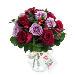 In this intimate bouquet, deep crimson and burgundy roses lean into one another like old friends, while pale lavender and dusty mauve roses offer a cool, calming counterpoint. Each rose is shown at full bloom: the petals are layered, slightly ruffled at the edges, and catch light in a way that reveals a satiny sheen. Bright red-orange hips are thoughtfully scattered through the mass of flowers, their small, rounded forms adding a lively, almost whimsical texture against the smoother rose petals. The dark green foliage - a backdrop of Ruscus or similar glossy leaves - pushes the colours forward, grounding the arrangement with a natural, leafy frame.

The bouquet is presented in a bulbous glass vase with a crackled, mercury finish that gives the piece a vintage, reflective quality; the vase narrows at the top and is finished with a piece of natural twine tied around the neck and a rectangular white tag bearing a simple rose illustration and flowing green lettering. The reflective surface of the vase catches soft, diffuse light and throws gentle highlights back into the blooms, enhancing the sense of depth and freshness.

I imagine this arrangement placed on a kitchen table in Clerkenwell as morning light strolls in from St. John Street, or wrapped and set for collection from a nearby shop on Hatton Garden. It feels tender and refined - an apt gift for saying thank-you, offering reassurance, or marking a modest celebration with elegance and warmth.
