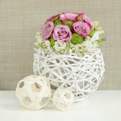 A romantic tabletop floral design is captured against a soft, linen-like beige background, evoking the relaxed elegance of a Clerkenwell wedding breakfast in one of the converted warehouses off St John's Square. In the centre of the image, a rounded bouquet rises from a white, woven rattan-style vase shaped like a slightly flattened sphere. The vase's open, criss-crossing strands create a lattice of light and shadow that feels both airy and hand-crafted, as if it had been woven especially for the occasion. Above it, roses in blush pink and gentle lavender unfold in generous blooms, their petal edges touched with a deeper, almost raspberry tone that adds depth and romance. Between the roses sit vivid green button chrysanthemums, whose small, domed heads give the arrangement energy and texture, like little bursts of spring foliage. Around the base of the bouquet, clusters of small white blossoms-dainty filler flowers with soft, rounded petals-peek out, creating a ruffled border that makes the design feel lush yet still perfectly controlled. The entire piece rests on a pristine white tabletop, its simplicity allowing the colours of the flowers to stand out clearly. To the left, two decorative wicker spheres in light, natural tones echo the vase's woven structure, bringing balance and a sense of thoughtful styling, reminiscent of a carefully dressed wedding table along Farringdon Road. The lighting is gentle and even, revealing every curve of petal and every twist of wicker, and the overall mood is quietly celebratory, ideal for a centrepiece welcoming guests to a ceremony or reception in Clerkenwell.
