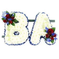 A tender, thoughtfully arranged floral tribute spelling BA, conceived for a memorial setting where craftsmanship and compassion meet. The letters are entirely faced with compact clusters of small white chrysanthemum heads, packed so densely they create a soft, cloud-like texture that seems to absorb the room's quiet. Each letter's shape is carefully traced in a ruffled royal blue satin ribbon, the rich blue edge both defining the initials and lending a restrained dignity to the overall composition. The B carries a floral accent on its upper left curve: a grouping of deep red roses-their petals glossy and velvety, at stages from button to half-open-accented with vivid blue delphinium-like spikes that give a cool, uplifting lift to the colour story. The A mirrors this touch with a matching cluster at its lower right, tying the pair together visually and emotionally. Lush dark green foliage supports the accents, while pale green, trailing tendrils soften the structural lines and add a gentle sense of movement. The letters rest side-by-side on a low dark green base for a freestanding presentation suitable for placement at a service or beside a memorial photograph. In soft morning or chapel light the whites glow quietly, the blues read as steady, and the red roses remind viewers of love and respect. The arrangement is crafted with the tempered rhythm of a seasoned florist, intended to provide a quiet, personal punctuation to farewell rituals and to comfort those who gather to remember.