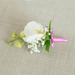 A refined orchid boutonniere is shown resting horizontally on a pale beige, linen-textured fabric, the kind of neutral backdrop that lets every floral detail shine, much like the understated elegance of Clerkenwell's townhouses and studios. At its heart sits a single, immaculate white Phalaenopsis orchid bloom, its broad, slightly cupped petals looking almost porcelain-smooth as they open to reveal a centre brushed with soft yellow and the faintest hint of blush pink. The orchid's throat is intricate yet gentle, drawing the eye inward without overwhelming the calm composition. Surrounding the base of this focal bloom, a halo of tiny white baby's breath flowers creates a misty, cloud-like ring, their countless small, round blossoms bringing lightness and air to the design. Slender, glossy green leaves tuck in behind the petals, giving a touch of freshness that suggests the boutonniere was crafted only moments ago in a local Clerkenwell flower shop. To one side, a short branching stem holds a pair of firm, lime-green orchid buds, tightly closed and smooth, evoking a sense of new beginnings and the anticipation of a wedding ceremony, prom, or elegant celebration. The stems are gathered with precision and bound in a vivid fuchsia pink satin ribbon, the bow tied neatly with a soft sheen that catches the light like a silk tie on a tailored suit. The colour pop of the ribbon contrasts beautifully with the angelic white orchid and airy gypsophila, making this floral accessory ideal for a groom or bridal party member arriving at a nearby Islington registry office or an intimate celebration just off Farringdon Road. The overall mood is one of composed romance and quiet sophistication, perfectly suited to modern city weddings.
