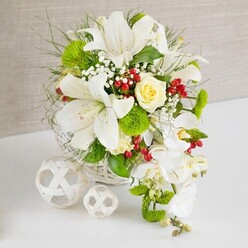 On a clean white tabletop, a luxurious wedding-style arrangement sits in a round white wicker basket, the composition quietly glowing against a soft beige, linen-like background reminiscent of a calm Clerkenwell studio. Several large white lilies form the upper structure, their wide, trumpet-shaped petals opening generously, each bloom revealing fine speckles and golden stamens that suggest a gentle, powdery scent. Cradled among them, a single pale cream rose, perfectly formed and slightly cupped, draws the eye to the heart of the design. Around this focal point, lush green textures create depth: bright green pom-pom chrysanthemums, possibly Santini or dianthus, protrude like soft spheres, while wisps of feathery foliage and polished deeper-green leaves fill in the spaces, giving the arrangement a natural, garden-fresh feel. Tiny sprays of white baby's breath thread through the flowers like light mist, softening every edge. Sparingly yet decisively placed, clusters of rich ruby-red berries provide a vivid contrast, adding a hint of winter warmth or festive vibrancy. Flowing from the right side, a cascade of elegant white phalaenopsis orchids drapes downward, their rounded petals and gentle curve lending a bridal, waterfall effect that would suit a ceremony at a Clerkenwell registry venue or an intimate celebration near St John Street. On the left of the composition, two decorative white woven balls rest on the tabletop, their airy latticework echoing the basket and reinforcing the modern, minimalist style. The overall impression is of a carefully balanced, serene floral centerpiece, perfect for wedding tables, anniversary dinners, or stylish gatherings in and around Clerkenwell.