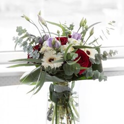 Illuminated by the gentle daylight that filters through Clerkenwell's distinctive Georgian windows, this floral composition strikes a graceful balance between classic charm and contemporary style. The focal point is a succession of sumptuous, deep red roses-their plush petals forming intricate spirals, reminiscent of conversations in Rosebery Avenue cafes. Elegant, creamy white gerberas with chocolate-brown centres lend their timeless appeal, standing shoulder to shoulder with airy white filler clusters that soften and expand the bouquet's visual presence. Artful touches of pale purple lisianthus thread through the blooms, some beginning to unfurl with a promise of joy, others retaining their budded mystery-a nod to blossoming moments. Generous sprays of fragrant eucalyptus arc outward, casting cool silver-green highlights and bringing a calming herbal note, akin to strolling through Spa Fields Park after a summer rain. Underpinning the ensemble, sculptural, dark green leaves are folded neatly, framing the arrangement and lending it gravitas. The stems, visible through a pristine glass vase, reveal the vibrant freshness emblematic of Clerkenwell's boutique florists. Ideal for thoughtful celebrations-whether adorning a Clerkenwell flat's kitchen table or conveying appreciation to a colleague-this bouquet's nuanced textures and aromas offer both sophistication and gentle solace, making it a treasure of the neighbourhood.