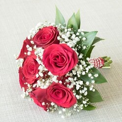 Nestled elegantly upon a pale, textured fabric-a linen reminiscent of Clerkenwell's creative studio districts-this enchanting bridesmaid bouquet is a celebration of classic romance, expertly hand-tied by local florists. At its core, several sumptuous red roses burst forth in full bloom, their petals tightly curled and richly layered in a swirl of velvety crimson. Between these iconic blooms, misty clouds of delicate white baby's breath float effortlessly, offering a dreamy, almost ethereal contrast that mirrors mist lingering over Farringdon rooftops. Dark green leaves, likely ruscus or glossy salal, weave through the bouquet, imparting a lush freshness that echoes the urban gardens dotted throughout Clerkenwell. The base of the bouquet is tightly bound with a striking ribbon in red and white stripes, its finish enhanced by a subtle glimmer of tiny white pearls-an elegant tribute to local weddings and milestone celebrations. Crafted with meticulous attention to detail, the arrangement's visual harmony is matched by the soft, inviting light, showcasing each petal, pearl, and leaf. This bouquet evokes a feeling of joyful celebration and sincere affection, suitable for a Clerkenwell wedding, anniversary, or any special moment shared in the heart of London. Its delicate scent and vibrant colours promise to enhance the atmosphere, carrying with it the spirit of skilled local craft and heartfelt tradition.