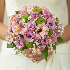 In Clerkenwell's charming surrounds, this bridal bouquet embodies elegance and a fresh sense of modern love. The bride's gentle hands encircle a lush, round collection of flowers, the subtle texture of white lace from her gown softly blurred in the background-reminiscent of cherished ceremonies along St John Street. Mauve and blush-pink roses sit at the heart of the arrangement, their delicate fragrance conjuring memories of early morning walks through Spa Fields' gardens. These timeless blooms, with petals at varying degrees of openness, are beautifully complemented by crisp lime green button chrysanthemums. Their spherical shape stands apart, adding a contemporary edge that mirrors Clerkenwell's blend of old and new. Blushing alstroemeria-each petal streaked with hints of maroon and sunlit yellow-lend the bouquet an exotic, graceful allure, while supple sprays of baby's breath fill the gaps with gentle softness and a hint of whimsy. Rich, dark green leaves peek through, anchoring the design in lush natural beauty. Every detail, from the light catching the bouquet to the tactile then visual contrast of petals and pom-pom mums, speaks to the artistry of local floristry. Here is a bouquet that could travel from a Registry Office on Rosebery Avenue to a bustling reception at a Clerkenwell restaurant-crafted as a joyful, fragrant symbol of union and new beginnings.