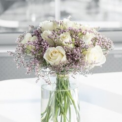 A refined, round bouquet of creamy white roses and soft lavender statice is captured in the centre of the frame, as though it has just arrived on a bright table in a Clerkenwell townhouse. The main impression is one of balance and stillness: around a dozen pristine white roses, their petals plush and gently curved, form a perfectly rounded crown. Each rose is at that ideal stage of opening, neither tight nor fully blown, so the inner whorls of petals are visible like carefully folded silk. Their colour is a warm cream that leans toward ivory, lending the whole bouquet a sense of quiet luxury. Between these generous blooms, threads of lilac statice create a fine, lace-like network, each diminutive flower giving a papery, slightly wild texture. The lavender tones remain delicate and understated, adding a soft veil of colour that never competes with the roses, instead enhancing their purity and depth. The stems are gathered in a tall, cylindrical glass vase, the clear sides revealing straight green stems and the clean line where they disappear into fresh water. Placed on a smooth, white surface, the arrangement seems to float in natural light pouring from an unseen window, similar to the gentle daylight that floods the creative studios and lofts around Clerkenwell Road and St John Street. The background falls away into a calm blur, so all attention rests on the flowers' subtle glow and the tiny shadows cast by each petal. The mood is composed and contemporary, suitable for a minimalist flat, a quiet breakfast table, or a thoughtful delivery to a friend working in one of Clerkenwell's design agencies-a bouquet that speaks softly of affection, remembrance, or heartfelt congratulations without a single word.