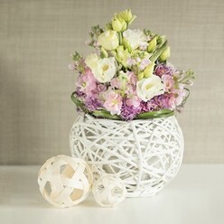 Placed elegantly in a sunlit Clerkenwell interior, this lush floral arrangement balances natural charm with contemporary designer flair, reminiscent of the neighbourhood's blend of history and modern aesthetics. The bouquet's heart comprises creamy white and gentle yellow lisianthus blooms, their velvety, ruffled petals layered for soft movement. Scattered among them, slender pink matthiola stocks reach upward, injecting vertical lift and subtle romantic energy. The base is enveloped in clusters of vibrant purple filler flowers-suggestive of wild asters found during Clerkenwell's spring strolls-offering vivid colour contrast and tactile interest. Encircling the blooms, deep green foliage neatly frames the arrangement, emphasizing freshness and clarity. Everything is presented in a rounded, open-work vase, crafted from white rattan in a web-like design that allows light to filter through, highlighting the artful composition within. To the arrangement's left, two light wood decorative spheres, echoing the vase's intricate weave, rest upon a bright white tabletop, their organic forms accenting the scene's artisanal quality. The whole is set before a soft, understated beige wall, lightly textured, reminiscent of the area's converted warehouse lofts and studio spaces. A testament to local craftsmanship and thoughtful design, this arrangement would make an ideal centrepiece for wedding celebrations, creative meetings, or intimate gatherings across Clerkenwell, infusing each setting with calm, freshness, and bespoke artistry from Clerkenwell Flowers.