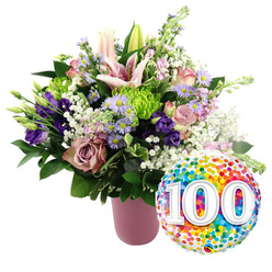 Set against a pure white background, this image captures a lavish centenary bouquet and balloon that feel as if they have just been arranged in a calm corner of a Clerkenwell studio, not far from Spa Fields and St John Street. A matte, ribbed pink vase anchors the composition on the left, its soft blush tone echoing the gentle romance of the flowers above. Rising from it, pale pink Oriental lilies stretch their star-like petals, some fully open to reveal pollen-dusted stamens, others still in tight, dignified buds that speak of days yet to be celebrated. Around these lilies nestle soft pink and muted mauve roses, their petals folding inwards like treasured memories. Deep violet lisianthus bloom between them, with velvety cups of colour that add drama and depth. Sprinkled throughout are lime green button chrysanthemums, perfectly rounded and textured, playing against airy sprays of white baby's breath that create a veil of lightness. Delicate lavender asters with bright yellow centres peep through the foliage, bringing a gentle countryside charm into this carefully structured, urban bouquet. Sleek dark green leaves and varied foliage frame the flowers, giving the design a full, lush silhouette while keeping the palette balanced and fresh. To the lower right, a round foil balloon overlaps the arrangement, its mirrored surface brimming with vivid confetti dots in reds, yellows, blues, greens, and purples. Across its centre, large white numerals "100" are printed with a soft shadow, bold and joyful, honouring a once-in-a-lifetime milestone. The image evokes a sense of pride and affection, ideal for marking a 100th birthday or centenary event anywhere in Clerkenwell, from creative loft spaces to long-loved family homes.