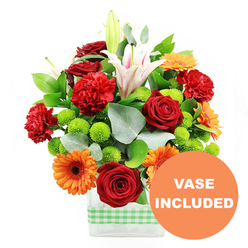 A lively mixed bouquet sits proudly against a pure white backdrop, presented in a modern, clear glass vase encircled at the base with a neat band of fresh light green and white gingham ribbon, reminiscent of picnic blankets in nearby Spa Fields. The flowers create a joyous, almost celebratory composition: velvety deep red roses and frilled red-orange carnations nestle side by side, their plush petals catching the light and giving the centre of the arrangement a warm, romantic glow. Bright orange gerbera daisies with dark, well-defined centres fan out around them, their flat, daisy-shaped faces adding a burst of optimism that feels perfectly in tune with Clerkenwell's creative energy. Scattered through the design are tight clusters of bright lime-green button chrysanthemums, their tiny, rounded heads adding a fresh, tactile contrast, like beads of colour. At the back and rising through the middle, elegant white Oriental lilies with soft pink accents lift the eye, some fully open with pollen-tipped stamens on display, others still wrapped in smooth buds, promising a slow, graceful unfurling over several days. The whole bouquet is framed with lush deep green foliage and long, slender stems of silvery eucalyptus that soften the outlines and add subtle movement, as though caught in a gentle breeze drifting up from Farringdon Road. In the lower right of the image, a round orange graphic partially overlaps the vase, clearly printed with the words "VASE INCLUDED" in bold white capitals, confirming that this contemporary glass container is part of the arrangement-ideal for quick, ready-to-display flower delivery to Clerkenwell apartments, studios, and offices.