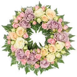 A beautifully composed circular funeral wreath fills the image, set against a clean white backdrop so that its colours and textures seem to float in quiet stillness. Every inch of the ring is covered with lush, fresh roses, creating a continuous band of petals that feels both soft and strong. Large, fully opened roses in creamy white and pale ivory provide a tranquil base, their generous blooms layered like folded silk. Nestled around them are clusters of blush pink spray roses, some still in bud, others just beginning to unfurl, hinting at gentleness and the lingering presence of love. Warm notes of peach and delicate buttery yellow roses glow softly within the composition, while distinctive dusty lavender and muted mauve roses introduce a thoughtful, slightly nostalgic tone, reminiscent of the historic brickwork and quiet side streets around Clerkenwell. Fine-textured, dark green fern foliage emerges between the flowers, adding depth, contrast, and a living, woodland freshness that grounds the pastel palette. The wreath forms a perfectly rounded circle, symbolising continuity, eternity, and the unbroken bond with the person being honoured. Light appears even and soft, with no harsh shadows, allowing the details of each petal and frond to be clearly visible, as if the arrangement has just been set down in a peaceful room before a service. This handcrafted tribute, suitable for memorials, funerals, or services at nearby chapels or crematoria serving Clerkenwell families, conveys quiet grace, compassion, and respect, offering a visually soothing focus for moments of reflection and farewell.