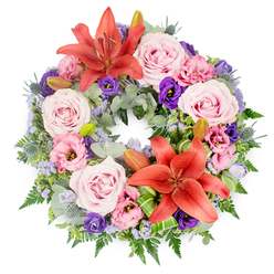 An abundant sympathy wreath lies in a perfect circle on a pure white background, its colours and forms carefully balanced to convey tenderness and quiet strength. A rich collar of greenery, including rounded eucalyptus leaves, feathery ferns, and other textured foliage, creates a deep green base that feels both natural and composed. From this foliage rise clusters of velvety blush-pink and soft lavender-pink roses, each one fully open and cupped, suggesting gentleness and care. Interlaced among them are lisianthus blooms, some in pale pink, others in a saturated purple that adds a thoughtful, contemplative note. Two bold Asiatic lilies in a vivid coral-orange, one near the top and one towards the lower section, draw the eye with their star-shaped petals and prominent stamens, their colour bringing a warm, hopeful light to the design. Several closed lily buds peek from the leaves, promising blooms that will unfold over the coming days. Spiky blue eryngium thistles and delicate sprays of lavender-blue statice offer cooler tones and fine, airy texture, like a soft haze hovering between the larger flowers. Carefully folded, striped green leaves are tucked around the lilies, giving the wreath a polished, contemporary finish. This floral tribute, crafted in the spirit of Clerkenwell's quiet side streets and historic churches, feels ideal for a funeral or remembrance service nearby, where families might gather to say goodbye. Its circular shape and layered pastels speak softly of continuity, cherished memories, and the enduring bond with a loved one who has passed.