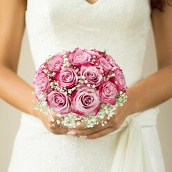The soft morning air in Clerkenwell seems to settle around this bouquet, where romance finds its form in each carefully chosen bloom. Close in the bride's hands is a round, almost jewel-like cluster of pink roses, their petals blending from a gentle blush at the core to an intense fuchsia along the ruffled margins. The effect is painterly, each rose revealing depth and character, much like the historic buildings along Clerkenwell Road. Interwoven are generous sprigs of feather-light white gypsophila, reminiscent of the mist that sometimes drifts along St John's Square at dawn. The bouquet is held just above the waist of a sleeveless dress, its bodice alive with delicate lace and an elegant V-shaped neckline. Subtle hints of a soft fabric sash gather to one side, adding another textural note. The bride's neatly manicured nails, painted pale and understated, rest atop the blooms - a detail as meticulously considered as every petal. In the softly blurred, neutral background, all the attention and drama belong to the floral arrangement, which balances exuberance with a sense of calm. This image reflects the soul of a Clerkenwell wedding: artistry, tradition, and an enduring promise, captured in flowers sourced and fashioned by artisans who know the heart of the area.
