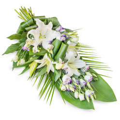 A refined sympathy spray is shown diagonally across a stark white backdrop, its softly tapering form evoking the gentle curve of a farewell gesture. The upper portion is full and rounded, where a cluster of white oriental lilies opens like quiet stars, their petals smooth and slightly curled at the tips, catching the light with a faint, satiny sheen. Some lilies remain as elongated buds, closed yet promising, standing among the open blooms like sentinels of memory. Woven through this focal point are graceful stems of lisianthus, their small, ruffled flowers predominantly white but kissed at the edges with lavender and lilac shading, as subtle as evening light over Exmouth Market. Tiny, unopened buds punctuate the arrangement, adding rhythm and depth. The flowers rest upon a generous foundation of foliage: wide, dark green leaves create a deep, protective cradle, while a few have been neatly folded and rolled into modern, sculptural accents that guide the eye along the length of the spray. From the right-hand side, slender palm-style fronds extend out in firm yet feathery arcs, giving the piece a sense of direction and flow, like a quiet walk from Farringdon up towards Clerkenwell Road. The palette is deliberately restrained-creamy whites, touches of lavender, and layered greens-so that the overall mood is one of serene contemplation rather than display. The arrangement feels cool and fresh, as though recently prepared in a local studio near St John's Square, designed to accompany a funeral, cremation, or memorial gathering with grace and heartfelt respect.