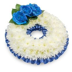 Captured from a slightly raised vantage that reveals its full circular silhouette, this memorial wreath is built from a thick, continuous band of immaculate white chrysanthemums, their layers of delicate, feathered petals forming a soft, textured surface that suggests serenity and repose. The chrysanthemum heads are closely set into a subtly domed profile; their tiny cream-tinted centres add a gentle warmth beneath the cool white. Anchoring the design at the upper left are three intense royal-blue roses, each petal velvety and precisely unfurled, planted into a bed of deep green foliage which sharpens their colour and lends the cluster presence without overpowering the whole. Around both the inner and outer perimeters a pleated satin ribbon in matching cobalt blue is neatly ruffled and secured, its glossy surface reflecting the studio light with restrained sparkle and lending structure to the composition. The wreath sits on a pristine white plane, a soft shadow beneath it giving depth and weight, and the arrangement's modest fragrance - the soothing, slightly spicy scent of fresh chrysanthemums - is implied rather than intrusive. As a Clerkenwell-based florist I imagine this piece quietly placed near Clerkenwell Green or delivered to a chapel on St John Street; its palette and craftsmanship make it especially fitting for services where dignity and tenderness are called for. The overall effect is one of calm refinement: traditional in material and meaning, but personalised through the dignified blue accents that suggest compassionate remembrance.