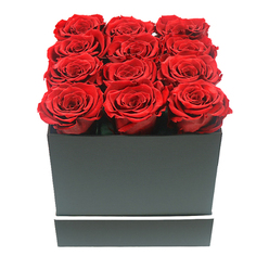 This close-up image presents an intimate, almost tactile view of a modern rose arrangement that feels perfectly at home in Clerkenwell's blend of history and design. At the centre of the composition sits a matte black square box, its edges crisp and architectural, reminiscent of the clean lines of local galleries and studios. Nestled inside are twelve sumptuous red roses, aligned in a meticulous three-by-four pattern so that each bloom gently brushes the next, forming a dense carpet of crimson. The roses appear fully open and expertly shaped, with tightly layered petals that spiral inwards to soft, shadowed centres, suggesting a preserved or long-lasting variety chosen for its ability to hold both form and colour. A narrow white band wraps horizontally around the base of the box, adding a refined, graphic detail that subtly echoes the monochrome signage seen around Clerkenwell's side streets. The background is pure white and uncluttered, isolating the floral design and making the red tones appear even deeper and more luxurious. Soft, balanced lighting illuminates every petal, bringing out the velvety texture and gentle curves, while faint shadows at the corners of the box provide depth without distraction. The mood conveyed is thoughtful and romantic rather than showy-an arrangement perfect for an anniversary dinner at home near Exmouth Market, a meaningful birthday gesture delivered to a creative studio off St John Street, or a quiet apology gift that speaks with sincerity. It is a contemporary expression of classic love, crafted with the attention to detail a local florist would bring to a special Clerkenwell delivery.